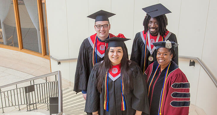 Four students in graduation caps and gowns by stairways.