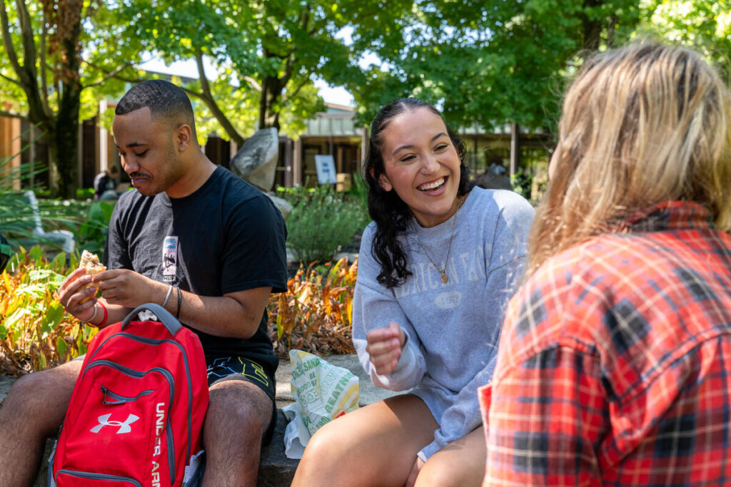 Three students outdoors talking and having lunch together.