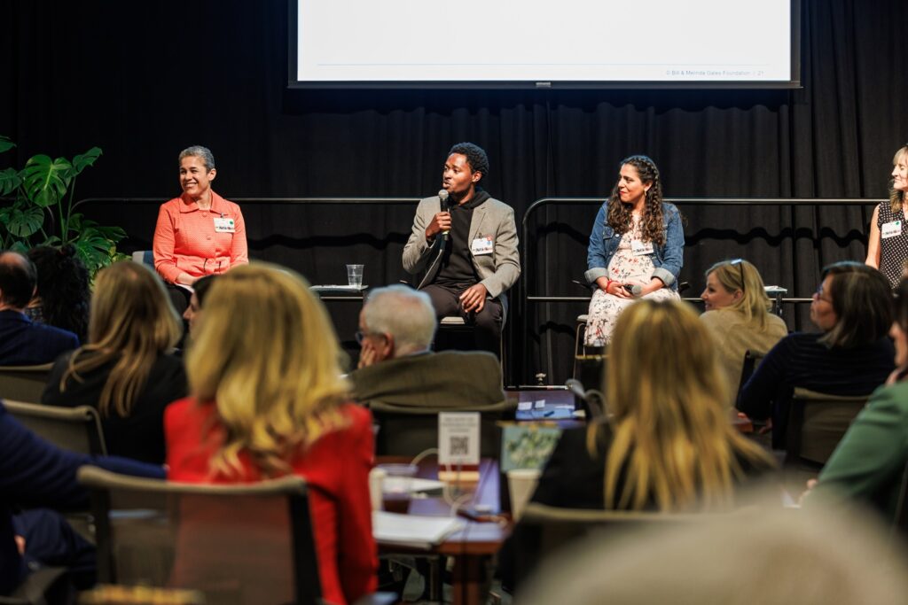 Four panelists holding microphones and sitting on chairs on a stage, one of them speaking to an audience